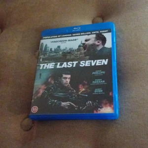 The Last Seven Blu-ray - Blu-ray av filmen 'The Last Seven' med ett dramatiskt omslag som visar en man med vapen och en stad i bakgrunden. Omslaget har en blå plastfodral och filmen är märkt med åldersgräns 15 år.