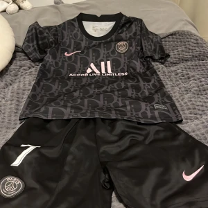 Svart PSG fotbollströja med Mbappé 7 - Snygg svart PSG fotbollströja från Nike med Mbappé och nummer 7 på ryggen. Tröjan har korta ärmar och ett diskret mönster i grått. Perfekt för fans av Paris Saint-Germain och Mbappé! 🖤