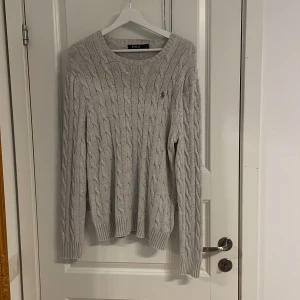 Grå stickad tröja från Polo Ralph Lauren - Säljer en stilren grå stickad tröja från Polo Ralph Lauren. Tröjan har ett klassiskt flätmönster och långa ärmar. Perfekt för kyligare dagar.