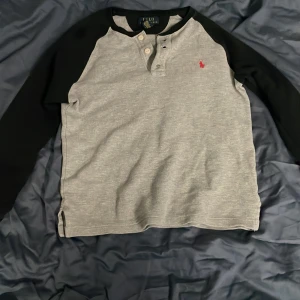 Grå och svart långärmad tröja från Polo Ralph Lauren - Säljer en stilren grå och svart långärmad tröja från Polo Ralph Lauren. Tröjan är i 100% bomull och har en knappdetalj vid halsen samt ett rött broderat märke på bröstet. Perfekt för en avslappnad stil.