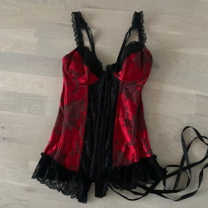 Röd och svart korsett från Shirley of Hollywood - Snygg röd och svart korsett med spetsdetaljer och snörning bak från Shirley of Hollywood. Den har justerbara axelband och en vacker blommig design. Perfekt för en elegant look.