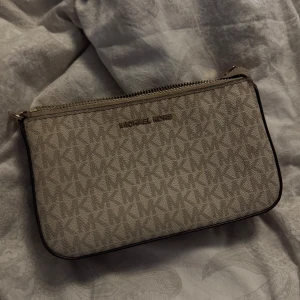 Beige kuvertväska från Michael Kors - Snygg beige kuvertväska från Michael Kors med kedjeaxelrem och dragkedja. Väskan har ett stilrent mönster med märkets logotyp och är perfekt för att bära det viktigaste. Den eleganta designen gör den lätt att matcha med olika outfits. det finns en del ”defekter” innuti väskan då den tyvärr är ” smutsig ” inget som syns utanpå väskan och annars i gott skick. går säkert att tvätta bort. 