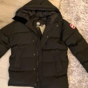  Canada Goose carson  - Säljer en svart dunjacka från Canada Goose med huva och dragkedja. Jackan har en klassisk design med varumärkets logga på ärmen. Perfekt för kalla vinterdagar.