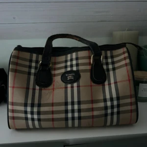  handväska från Burberry - Äkta men  slitna handtag. Snygg handväska från Burberry med klassiskt rutigt mönster i beige, svart och rött. Väskan har svarta handtag och detaljer i skinn. Perfekt för en stilren look.