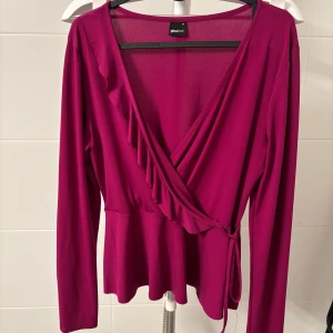 Lila top från Gina  - Snygg lila omlottopp från Gina med volangdetaljer och knytband i midjan. Toppen har långa ärmar och en v-ringad design som ger en elegant look. Nyskick 
