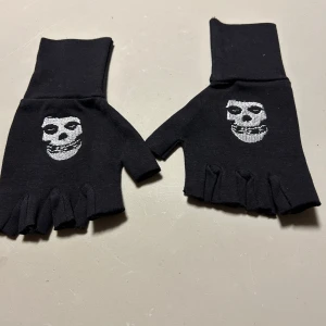 Svarta fingerlösa handskar med dödskallemotiv - Coola svarta fingerlösa handskar med ett vitt dödskallemotiv på ovansidan. Perfekta för att ge en edgy touch till din outfit. De är elastiska och bekväma att bära. Miss fits.