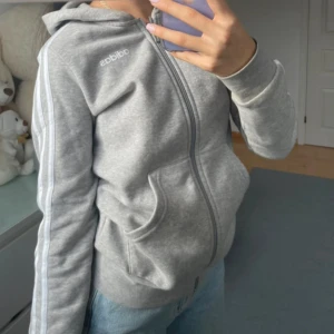 Grå huvtröja från Adidas - Säljer en grå huvtröja från Adidas med dragkedja och klassiska ränder längs ärmarna. Helt ny doxk utan prislapp