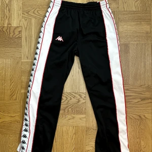 Svarta byxor från Kappa - Snygga svarta träningsbyxor från Kappa med vita sidopaneler och logotyper längs benen. Byxorna har elastisk midja och knappar längs sidorna för justerbar passform. Perfekta för en sportig look:) storlek passar S/M