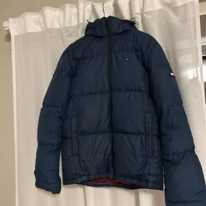 Blå pufferjacka från Hilfiger Denim - Säljer en snygg blå pufferjacka från Hilfiger Denim. Jackan har en dragkedja framtill och en praktisk huva. Den har gått sönder (men blivit lagad) dels på vänster arm och liknande märke vid midjan