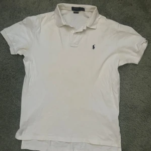 Vit pikétröja från Ralph Lauren - Klassisk vit pikétröja från Ralph Lauren med broderad logga på bröstet. Tröjan har korta ärmar och en stilren krage med knappar. Perfekt för en avslappnad och stilren look.