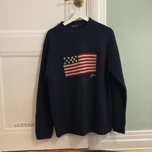 Mörkblå stickad tröja från Gant - Säljer en mörkblå stickad tröja från Gant med ett amerikanskt flaggmotiv på framsidan. Tröjan har långa ärmar och en rund halsringning. Perfekt för en avslappnad stil.