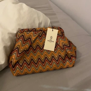 Mönstrad väska från Beck Söndergaard - Snygg väska från Beck Söndergaard med ett färgglatt sicksackmönster i orange, rött och gult. Perfekt för att ge din outfit en färgklick. Väskan har en praktisk storlek och är lätt att bära med sig.