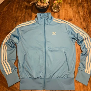 Blå träningsjacka från Adidas - Snygg ljusblå träningsjacka från Adidas med klassiska vita ränder längs ärmarna. Jackan har dragkedja och hög krage, perfekt för en sportig look. Adidas-loggan är broderad på bröstet. Passar perfekt för både träning och avslappnade dagar.