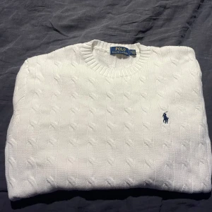 Ralph Lauren || Cable Knit Cotton - Säljer en klassisk vit kabelstickad tröja från Polo Ralph Lauren. Tröjan har en rund halsringning och det ikoniska logotypbroderiet i blått på bröstet. Perfekt för en stilren look. Kvitto finns 