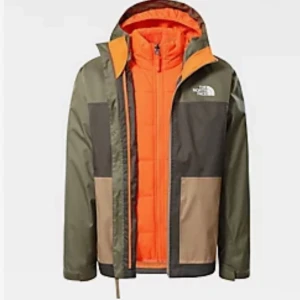 Flerfärgad jacka från The North Face - Snygg flerfärgad skid jacka från The North Face i grönt, brunt och beige med orange foder. Innerfodret är även avtagbart om man vill det. Jackan har en dragkedja och huva, perfekt för skidåkning och utomhusaktiviteter. Den har långa ärmar och praktiska fickor framtill.