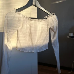Vit offshoulder topp - Säljer en stilren vit offshoulder topp med långa ärmar. Toppen är croppad och har en ribbad struktur som ger en snygg passform. Perfekt för både vardag och festliga tillfällen. Använd en gång, säljer då den aldrig använts igen! 