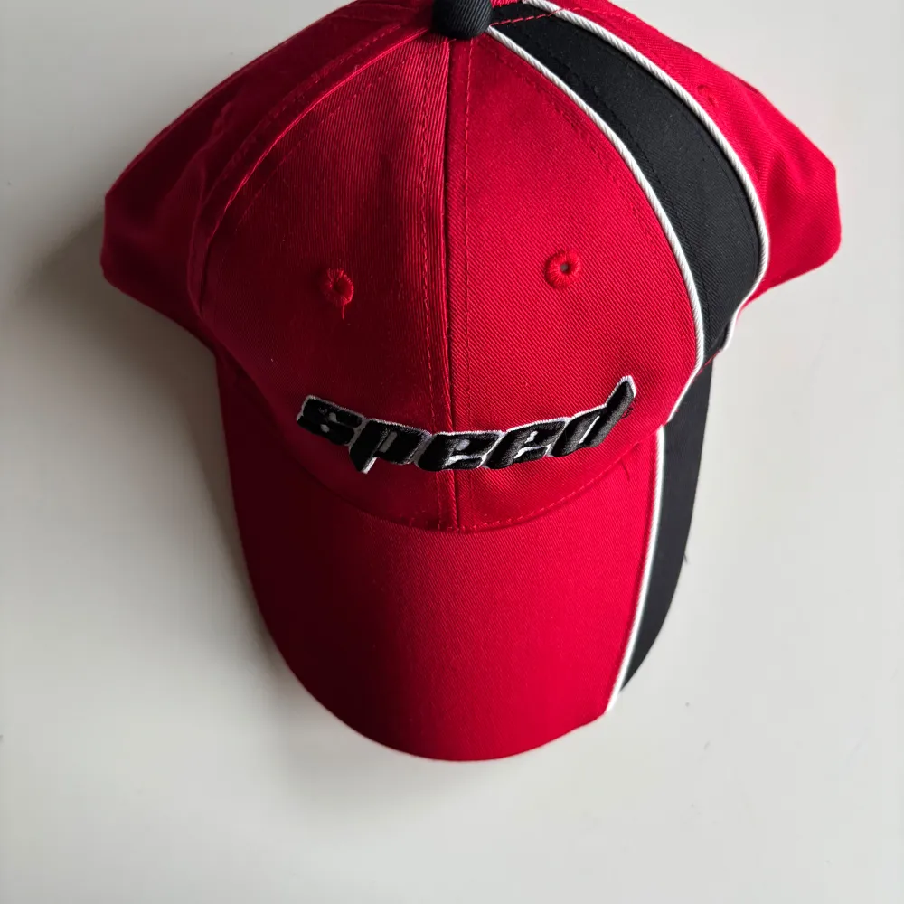 Snygg röd keps med svart och vit detalj på sidan och texten 'speed' broderad framtill. Perfekt för en sportig look.. Asusteet.