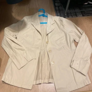 Beige kavaj med knappar - Säljer en stilren och mångsidig beige jacka i utmärkt skick! Denna jacka är perfekt för både vardagsbruk och mer formella tillfällen. Den har en klassisk design med en enkel knappslutning och är gjord av bekvämt och lätt material som passar bra för alla säsonger.   Ge din garderob ett lyft med denna tidlösa jacka! Perfekt att styla med jeans eller klänning. Missa inte chansen att förvärva detta plagg!