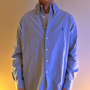Blå skjorta från Ralph Lauren - Snygg blå skjorta från Ralph Lauren med klassisk krage och knappar framtill. Den har en broderad logga på bröstet. Perfekt för en stilren look.