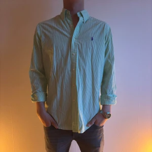 Grön- och vitrandig skjorta från Ralph Lauren - Snygg grön- och vitrandig skjorta från Ralph Lauren med klassisk krage. Skjortan har en broderad logga på bröstet och knappar framtill. Perfekt för en stilren look.