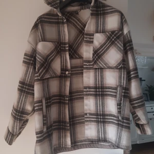Rutig overshirt i beige och svart - Säljer en snygg rutig overshirt i beige och svart med knappar framtill. Jackan har långa ärmar och två stora bröstfickor. Perfekt för våren som är påväg.
