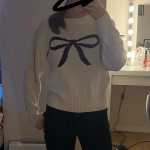 Vit stickad tröja med rosett - Säljer en söt vit stickad tröja med en stor svart rosett på framsidan. Tröjan har långa ärmar och ribbade kanter vid ärmslut och nederkant. Perfekt för en mysig dag!