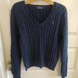 Mörkblå stickad tröja från Polo Ralph Lauren - Säljer denna stilrena kabelstickade v-ringade tröjan från Ralph Lauren, marinblå med vinröd logga. Stl M, borde även funka för en S beroende på hur du vill att den skall sitta, se mått. Mycket fint skick, inga hål eller fläckar, mjukt och stretchig kvalitet. Mått,ostretchat: Armhåla till armhåla 45cm, bredd längst ner 37cm, längd från mitt på axeln 56cm.