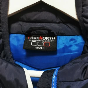 Svart pufferjacka från True North - Säljer en svart pufferjacka från True North, perfekt för kyliga dagar. Jackan har en dragkedja i blått och är quiltad för extra värme. Den är designad för nordiskt väder och har en stilren look.