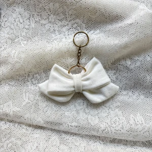 Vit rosett i tyg - Charmig vit rosett med en elegant design. Perfekt som en söt accessoar till väskan eller nycklarna. Rosetten är fäst vid en guldfärgad kedja och ring.