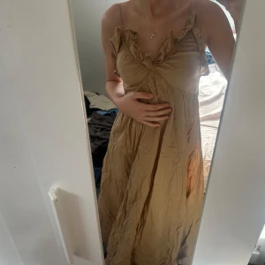 Beige klänning med volangdetaljer - Charmig beige klänning med volangdetaljer vid axlarna och en vacker omlott-design framtill. Klänningen har smala axelband och en luftig passform som ger en romantisk känsla.