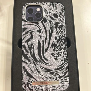 Hypnotic Sparkle mobilskal från iDeal of Sweden - Snyggt mobilskal för iPhone 12 Pro Max med ett hypnotiskt glittrigt mönster i svart och silver. Perfekt för att ge din telefon en stilfull och unik look. Skalet är både trendigt och skyddande.