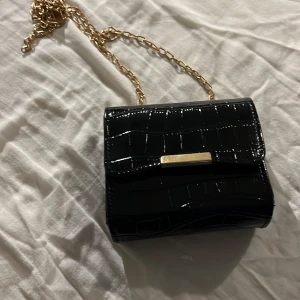 Svart axelväska med kedja - Snygg svart axelväska och guldfärgad kedja. Väskan har en elegant design med en metallplatta på framsidan. Perfekt för att ge en stilren touch till din outfit.