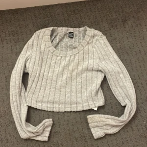 Grå ribbad croppad tröja - Säljer en stilren grå ribbad croppad tröja med långa ärmar. Perfekt för en avslappnad look. Tröjan har en rund halsringning och är i en mjuk och bekväm kvalitet.