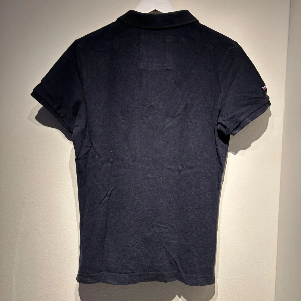 Snygg svart pikétröja från Superdry med korta ärmar och klassisk krage. Tröjan har en liten broderad logga på bröstet och knappar framtill. Perfekt för en stilren look.. T-paidat.