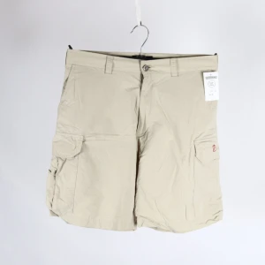 Beige shorts från Fjällräven - Säljer ett par beige shorts från Fjällräven med praktiska fickor och knappstängning. Tillverkade i en slitstark blandning av polyamid och bomull, perfekta för utomhusaktiviteter. Klassisk design med bälteshällor och en bekväm passform. Köpta secondhand, myrornas prislapp är kvar och är på 450 kr, mitt pris är 300 kr.