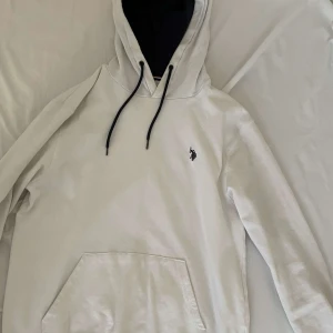 Vit hoodie från U.S. Polo Assn. - Säljer en stilren vit hoodie från U.S. Polo Assn. med svart logga på bröstet. Hoodien har en justerbar huva med svarta snören och en praktisk magficka. Perfekt för en avslappnad stil. Den är helt ny och skönt använda passa me allt stil