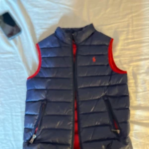 Blå dunväst från Ralph Lauren - Säljer en snygg blå dunväst från Ralph Lauren med röd insida och detaljer. Västen har en dragkedja framtill och är perfekt för kyligare dagar. Den ikoniska loggan syns på bröstet. Passar bra för en stilren look. Västen är junior size!