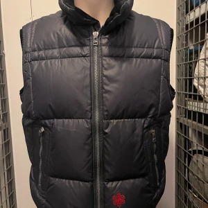 Grå dunväst från J.Lindeberg - Säljer en grå dunväst från J.Lindeberg med dragkedja och två fickor fram. Västen har en quiltad design och är fodrad med röd insida. Perfekt för kyliga dagar.