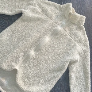 Krämvit teddyjacka från Ginatricot  - Mysig vit teddyjacka från Smartwool med dragkedja framtill. Perfekt för kyliga dagar med sin fluffiga och varma design. Jackan har en hög krage och långa ärmar för extra värme.