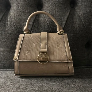 Beige handväska, juice couture  - Snygg beige handväska med präglad logotyp och guldfärgad detalj framtill. Väskan har ett handtag och en avtagbar axelrem med kedjedetaljer. Perfekt för att ge din outfit en elegant touch.