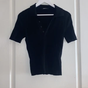 Svart ribbad pikétröja från Gina Tricot - Snygg svart ribbad pikétröja från Gina Tricot med korta ärmar och knappar framtill. Perfekt för en stilren look. Passar bra till både jeans och kjol.