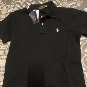 Svart pikétröja från Ralph Lauren - Snygg svart pikétröja från Ralph Lauren med klassisk krage och två knappar. Den har en broderad logotyp i vitt på bröstet. Perfekt för en stilren look.