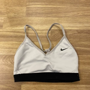 Vit sporttopp från Nike - Säljer en vit sporttopp från Nike med smala axelband och en svart resårkant nedtill. Toppen har en enkel design med en liten Nike-logga på framsidan. Perfekt för träning eller vardagsbruk. Lite nopprig 