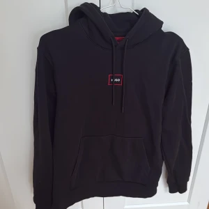 Svart hoodie från Hugo Boss - Säljer en stilren svart hoodie från Hugo med en röd logotyp på bröstet. Hoodien har en klassisk design med känguruficka och justerbar huva med snören. Perfekt för en avslappnad look. Använd en gång och tvättad en gång. Nypris 1400kr 