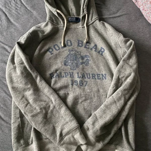 Grå hoodie från Ralph Lauren - Säljer en grå hoodie från Ralph Lauren med ett tryck av Polo Bear och texten 'Ralph Lauren 1967' i blått. Tröjan har en klassisk känguruficka och justerbar huva med snören. Perfekt för en avslappnad stil. Tar trades‼️‼️