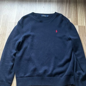 Marinblå tröja från Ralph Lauren - Säljer en stilren marinkblå tröja från Ralph Lauren med det klassiska röda logotypen på bröstet. Tröjan har långa ärmar och ribbade kanter vid ärmslut och nederkant. Perfekt för en elegant och avslappnad look.