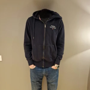 Mörkblå hoodie från Tommy Hilfiger - Säljer en snygg mörkblå hoodie från Tommy Hilfiger med dragkedja och broderad logga på bröstet. Den har långa ärmar och en bekväm passform. Vid minsta funderingar är det bara att höra av sig// Merch Åled 
