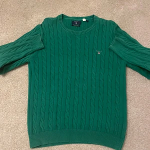 Grön kabelstickad tröja från GANT - Säljer en snygg grön kabelstickad tröja från GANT i premium bomull. Tröjan har en klassisk design med rund hals och långa ärmar. Perfekt för en stilren look. 💚
