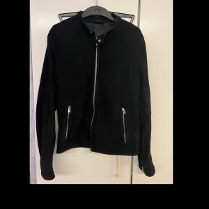 Svart mockajacka från Rock&Blue - Snygg svart mockajacka med dragkedja framtill och två dragkedjefickor. Jackan har en stilren design med långa ärmar och en hög krage. Perfekt för en cool och avslappnad look.