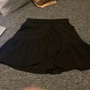 Svart omlottkjol - Snygg svart omlottkjol med volangdetaljer. Kjolen har en elastisk midja för bekväm passform och är perfekt för en stilren look. Den är kort och passar bra till olika tillfällen.